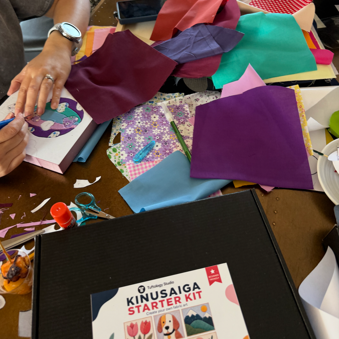 A black binder labeled 'KINUSAIGA STARTER KIT' sits on a cluttered craft table surrounded by colorful fabric pieces, scissors, and hands working on a project.
