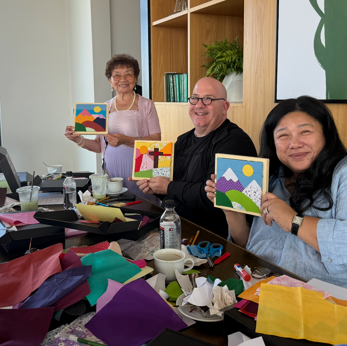 Kinusaiga Workshop Three people sitting and standing around a table filled with colorful craft supplies, each holding up a small painted mountain landscape artwork they created together.